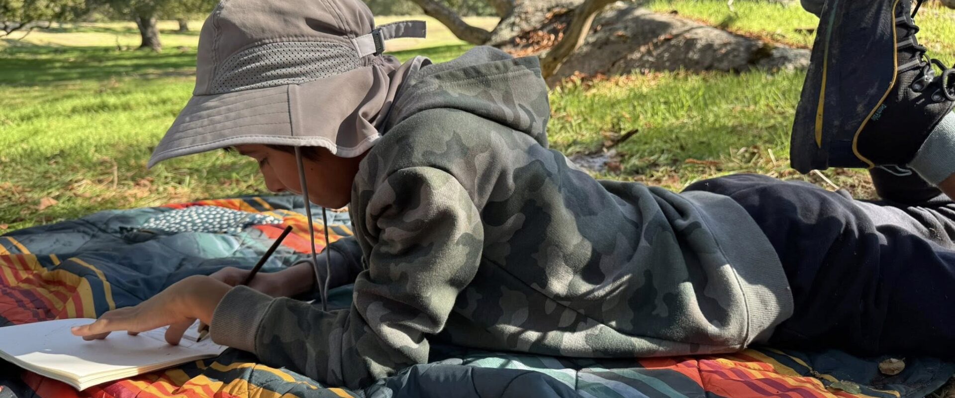Child journaling and drawing under oak trees with colored pencils